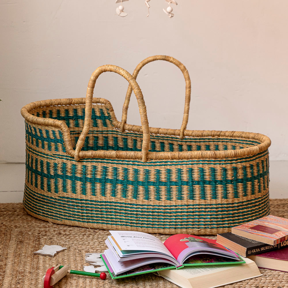 Moses basket with greenish stripes and long handles on a rattan rug surrounded by children’s books.  Designed by Akan Republic.